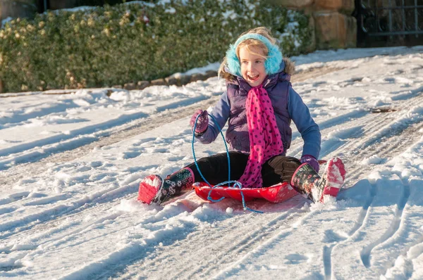 Laughing, vibrantly dressed young girl with blue ear muffs sledging down a slope — Stock Photo, Image