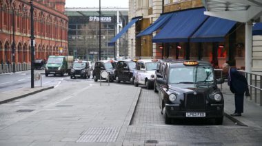 19 Mart 2020: Takım elbiseli yaşlı bir adam King 's Cross St. Pancras Tren İstasyonu' nun önünde bekleyen siyah bir Londra taksisine biniyor ve Coronavirus salgını sırasında PPE maskeli gençler yanından geçiyor.