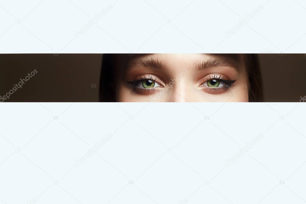 beautiful eyes peep into the crack. young beautiful woman. make-up artist concept. A girl with beautiful bright green eyes with shining shadows, looks into the hole of white paper