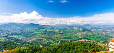 Vadi, yeşil tepeler, tarlalar ve mavi gökyüzü beyaz bulutlarıyla kaplı San Marino banliyö bölgesinin köylerinin panoramik manzarası. San Marino kalesinden görüntü.