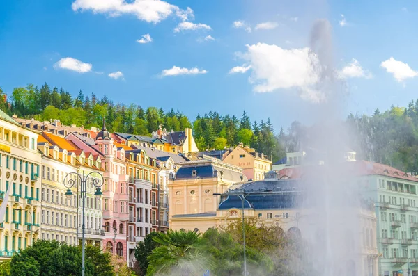 Sıcak kaynak gayzer Vridlo Karlovy Vary Carlsbad tarihi şehir merkezinde, renkli güzel binalar, mavi gökyüzü beyaz bulutlar, yeşil ağaçlı orman, Batı Bohemya, Çek Cumhuriyeti