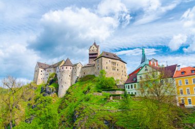 Madalyon Şatosu Hrad Loket Gotik tarzı binalar, Loket kasabasındaki devasa kaya ve renkli binalar, yeşil ağaçlar ve tepeler, mavi bulutlu gökyüzü arka planı, Karlovy Vary Bölgesi, Batı Bohemya, Çek Cumhuriyeti