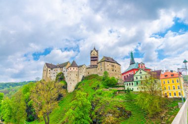 Madalyon Şatosu Hrad Loket Gotik tarzı binalar, Loket kasabasındaki devasa kaya ve renkli binalar, yeşil ağaçlar ve tepeler, mavi bulutlu gökyüzü arka planı, Karlovy Vary Bölgesi, Batı Bohemya, Çek Cumhuriyeti