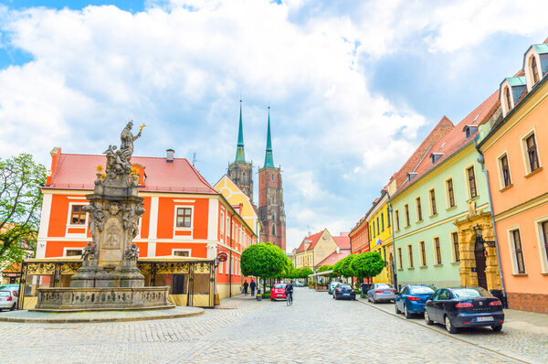 Wroclaw, Poland, May 7, 2019: Monument on square and cobblestone road street with green trees to Cathedral of St. John the Baptist building with two spires in old historical city centre, Ostrow Tumski