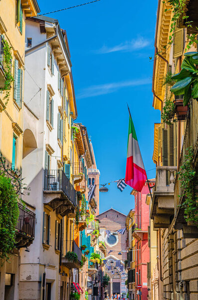 Typical italian street with traditional colorful buildings with shutter windows, balconies plants and flag, Basilica di Santa Anastasia church, Verona city historical centre, Veneto Region, Italy