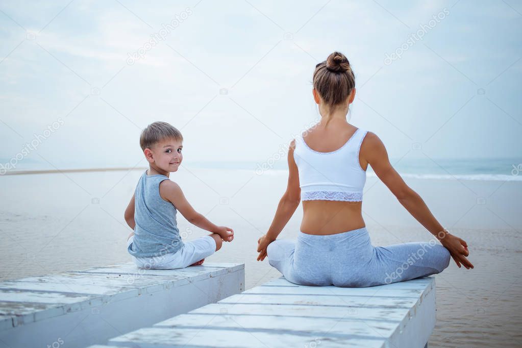 A mother and a son are doing yoga exercises at the seashore of t