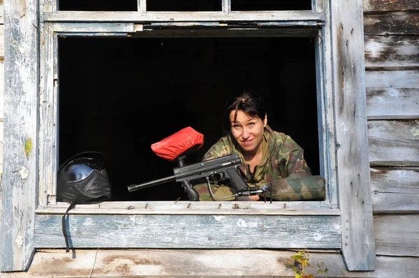 Молодая девушка в камуфляже рядом с защитной маской и с пейнтбольным маркером в окне заброшенного здания . — стоковое фото
