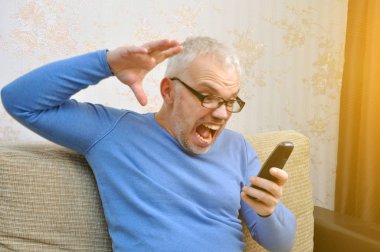 Mavi kazaklı, genç ve sağlıklı bir adam evde kanepede otururken telefonla konuşuyor..