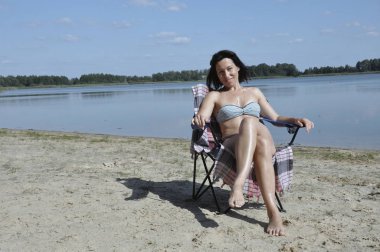 Genç çekici kadın bir sandalyede oturuyor ve göl kenarında güzel bir manzarası olan doğada dinleniyor..