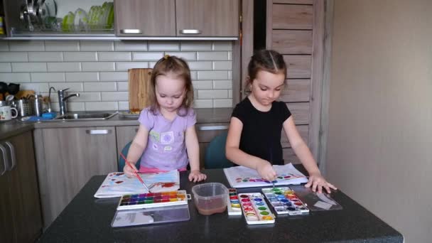 İki küçük kız kardeş mutfaktaki bir masada çocuklarla resim yapıyor. — Stok video