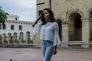 Sokakta poz veren 9-25 yaşlarındaki güzel bir kızın açık hava portresi. Beyaz bluz, dar kot pantolon ve sapatilla giyiyor. Şehir yaşam tarzı. Kadın moda konsepti.