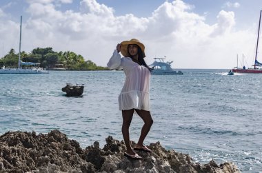 25-30 yaşları arasında siyahi bir kadın, kayaların üzerinde duruyor, beyaz mankenlik yapıyor, taze yaz ve tropikal kıyafetler giyiyor, plaj manzarası ve güneşli gökyüzü var.