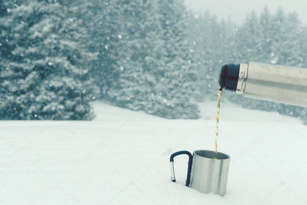 Tea from thermos pouring into a travel mug standing on the snow. Outdoors winter landscape background with snowy spruce