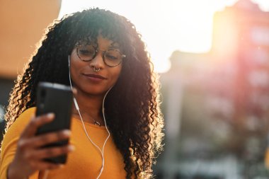 Kıvırcık saçlı, hareketli yaşam tarzı konseptiyle müzik dinleyen genç bir Afro-Amerikalı kadın.