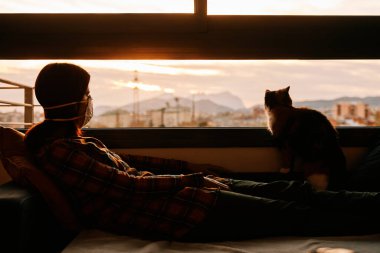 Maskeli genç bir kadın, Coronavirus krizi sırasında evde otururken geleceğe bakar.