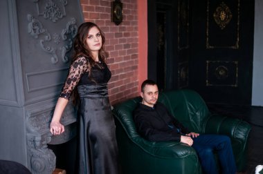 Sandalyede oturan adam ve duvarın yanında oturan kadın.