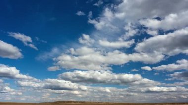 Zaman, rüzgâr türbinlerinin altındaki mavi gökyüzüne karşı beyaz kabarık bulutları eritiyor.