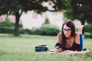 Genç bir kadın akıllı telefon kullanarak yaz parkında dinleniyor.