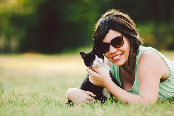 美しい若い女性が公園の芝生の上に横になって猫を抱いています — ストック写真