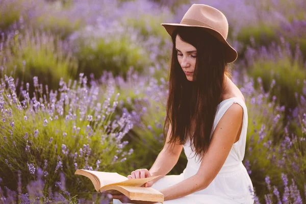 白いドレスを着た美しい女性は日没時に畑のラベンダーの花の間で本を読んで — ストック写真