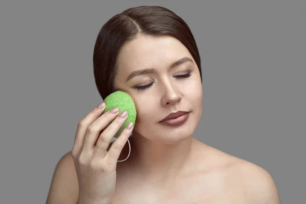 Kız yeşil bir konjac süngerle gri bir arka planda Tex için yer ile yüz tedavisi yapıyor.