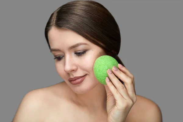 Kız yeşil bir konjac süngerle gri bir arka planda Tex için yer ile yüz tedavisi yapıyor.