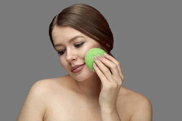Kız yeşil bir konjac süngerle gri bir arka planda Tex için yer ile yüz tedavisi yapıyor.
