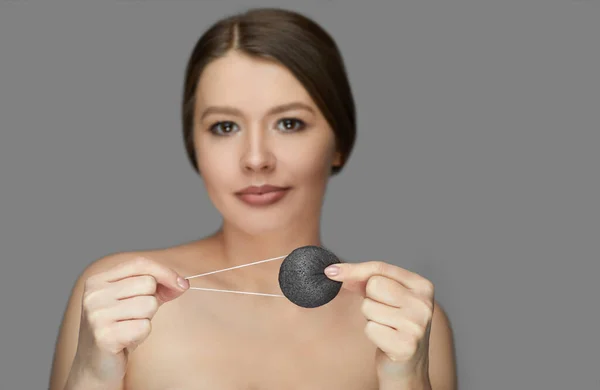 Kız gri arka planda siyah bir konjac süngerle yüz bakımı yapıyor.