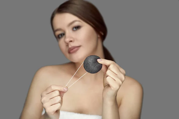 Kız gri arka planda siyah bir konjac süngerle yüz bakımı yapıyor.