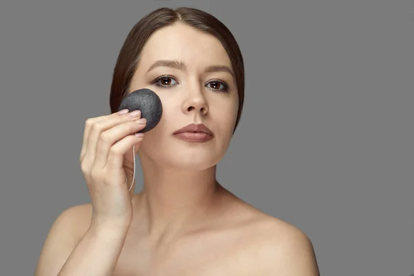 Kız gri arka planda siyah bir konjac süngerle yüz bakımı yapıyor.