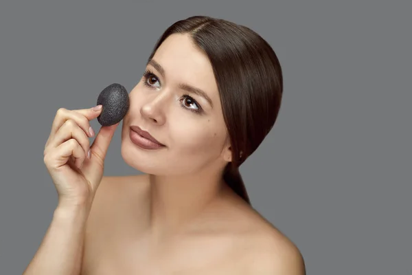 Kız gri arka planda siyah bir konjac süngerle yüz bakımı yapıyor.