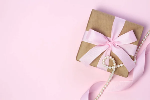 Pembe fiyonklu bir hediye kutusu ve pastel pembe arka planda inci boncuklar. Düzleştirme biçimi.