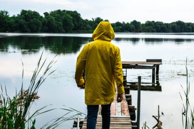 Sarı su geçirmez yağmurluklu şık genç adam gölün veya nehrin kıyısındaki ahşap iskelede duruyor.
