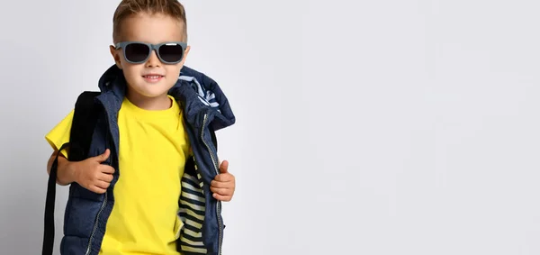 Attraktiv liten pojke i snygga varma kläder med en ryggsäck på axlarna, ha kul på en ljus studio bakgrund. — Stockfoto