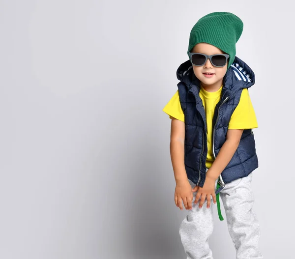 Стильный веселый мальчик 4 лет в свитерах, зеленой шляпе, очках и пуховом жилете веселится — стоковое фото
