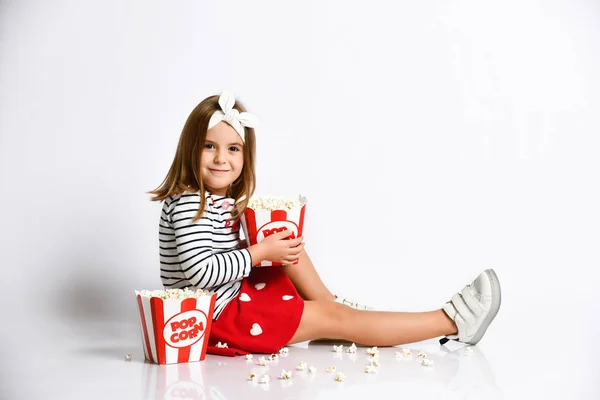 Молода дівчинка в червоній спідниці сидить на підлозі з відром попкорну на сірому фоні — стокове фото