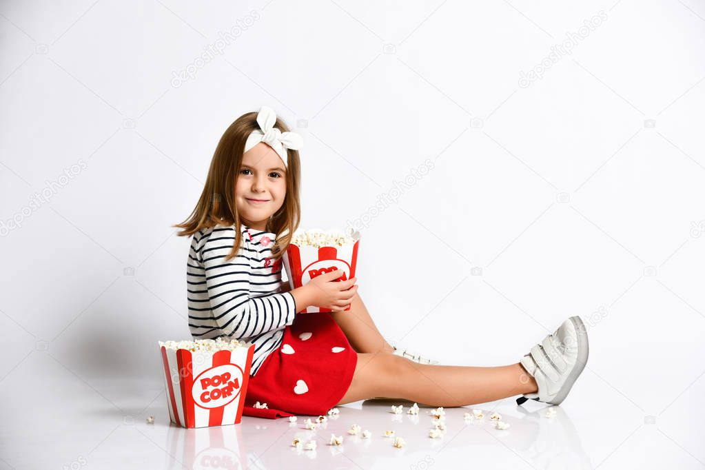 Young little girl in a red skirt sits on the floor with a bucket of popcorn on a gray background
