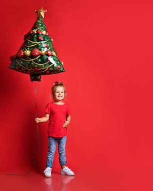 Kırmızı tişörtlü, mavi kotlu, kırmızı arka planda bedava fotokopi alanı olan bir Noel ağacı balonu tutan mutlu küçük kız..