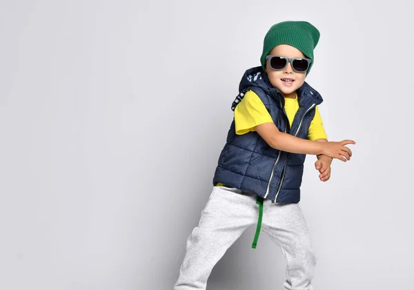 Elegante niño alegre 4 años de edad en pantalones de chándal, un sombrero verde, gafas y un chaleco abajo se divierte —  Fotos de Stock