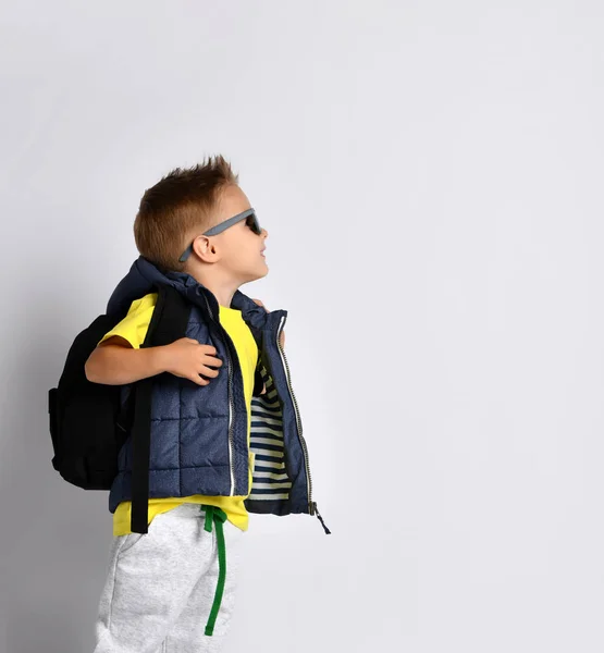 Atractivo niño en ropa de abrigo con estilo con una mochila en los hombros, divertirse en un fondo de estudio de luz . —  Fotos de Stock