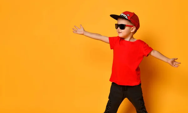 Ung kille pojke i en röd T-shirt och mörka byxor, vita sneakers och en rolig mössa poserar på en gratis kopia utrymme på en gul bakgrund — Stockfoto