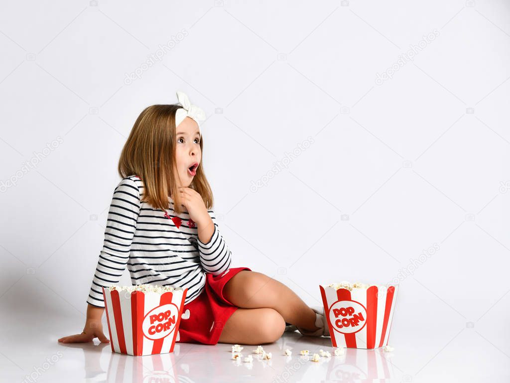 Surprised little girl in a red skirt sits on the floor with a bucket of popcorn on a gray background