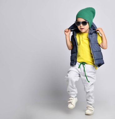 4 yaşında, eşofmanlı, yeşil şapkalı, gözlüklü ve yelekli bir çocuk eğleniyor.