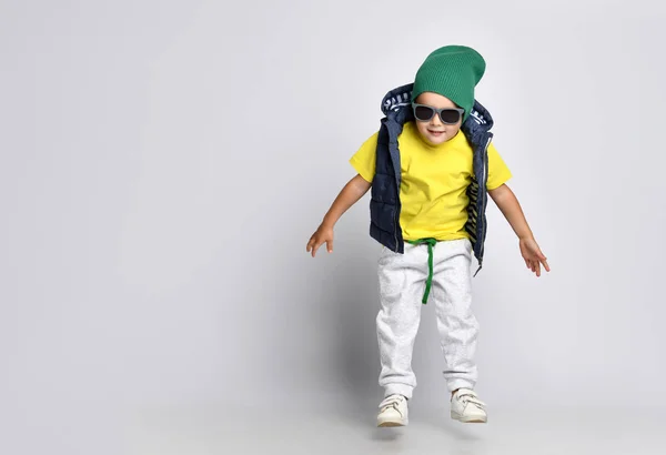 Snygg glad liten pojke 4 år gammal i sweatpants, en grön hatt, glasögon och en dunväst har kul — Stockfoto