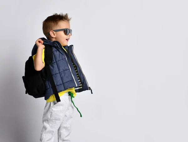 Attractive little boy in stylish warm clothes with a backpack on his shoulders, having fun on a light studio background. — Stock Photo, Image