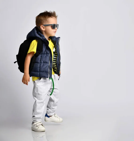 Привабливий маленький хлопчик у стильному теплому одязі з рюкзаком на плечах, розважається на світлому фоні студії . — стокове фото