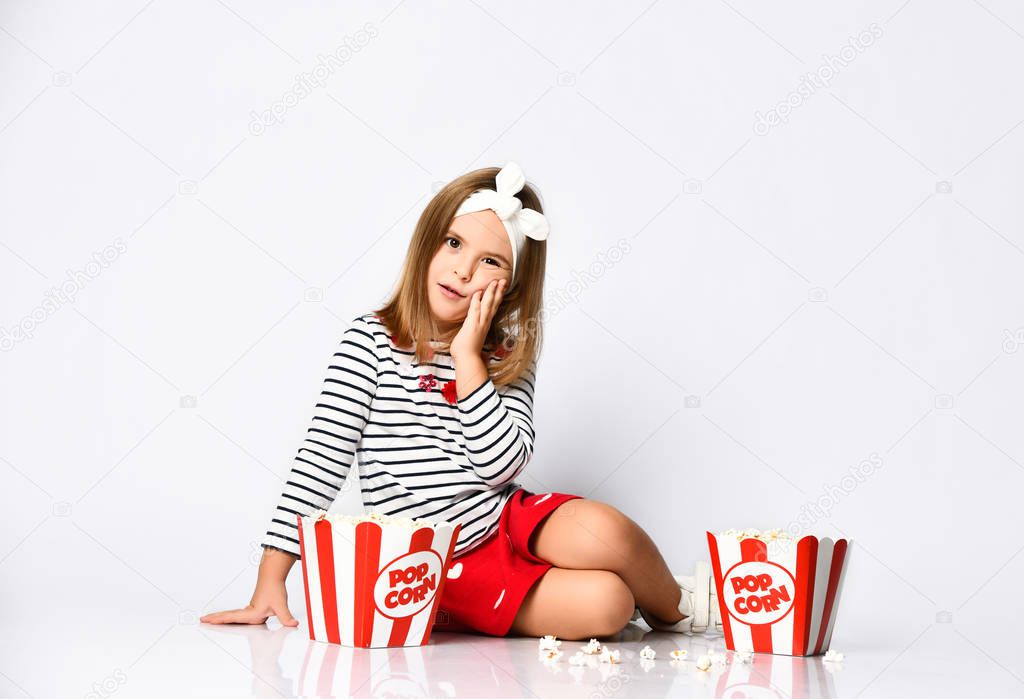 Young little girl in a red skirt sits on the floor with a bucket of popcorn on a gray background
