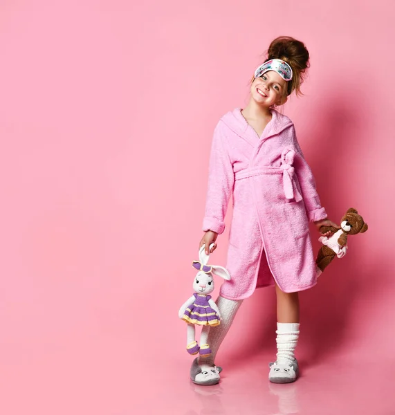 Grappig meisje in roze huisjurk en pluche sneakers houdt twee speelgoed — Stockfoto