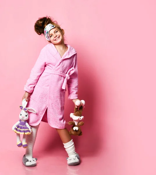 Grappig meisje in roze huisjurk en pluche sneakers houdt twee speelgoed — Stockfoto