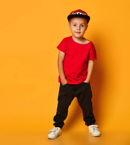 Ung kille pojke i en röd T-shirt och mörka byxor, vita sneakers och en rolig mössa poserar på en gratis kopia utrymme på en gul bakgrund — Stockfoto